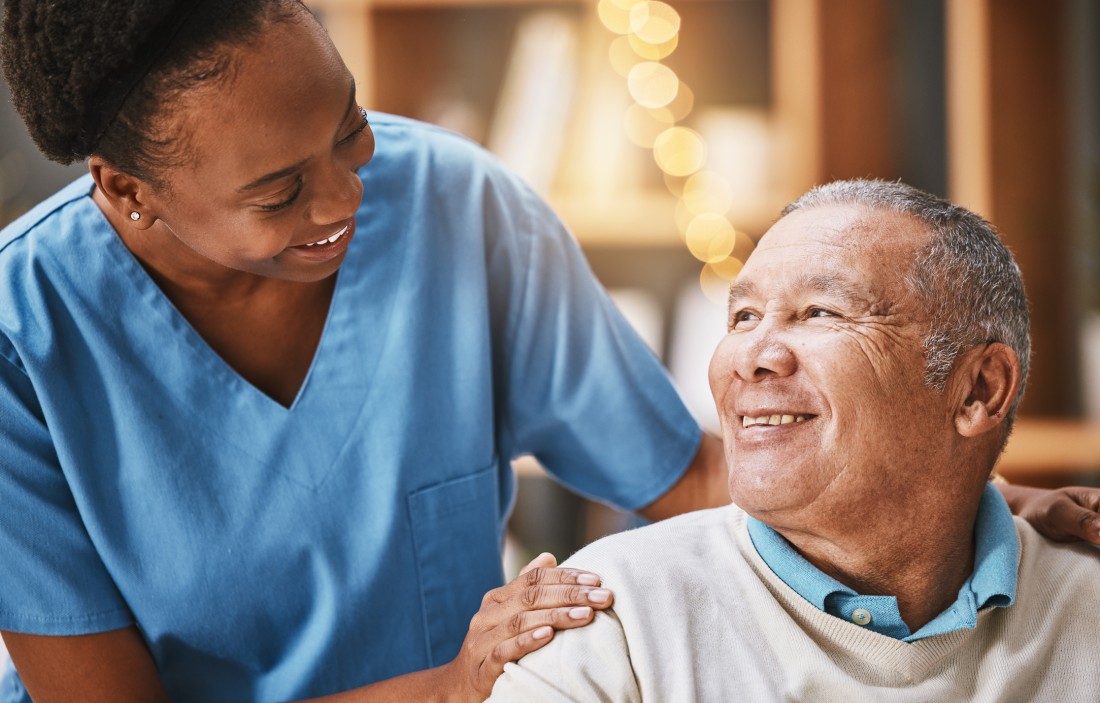 A home caregiver rests her hands on a senior client’s shoulders as they both smile