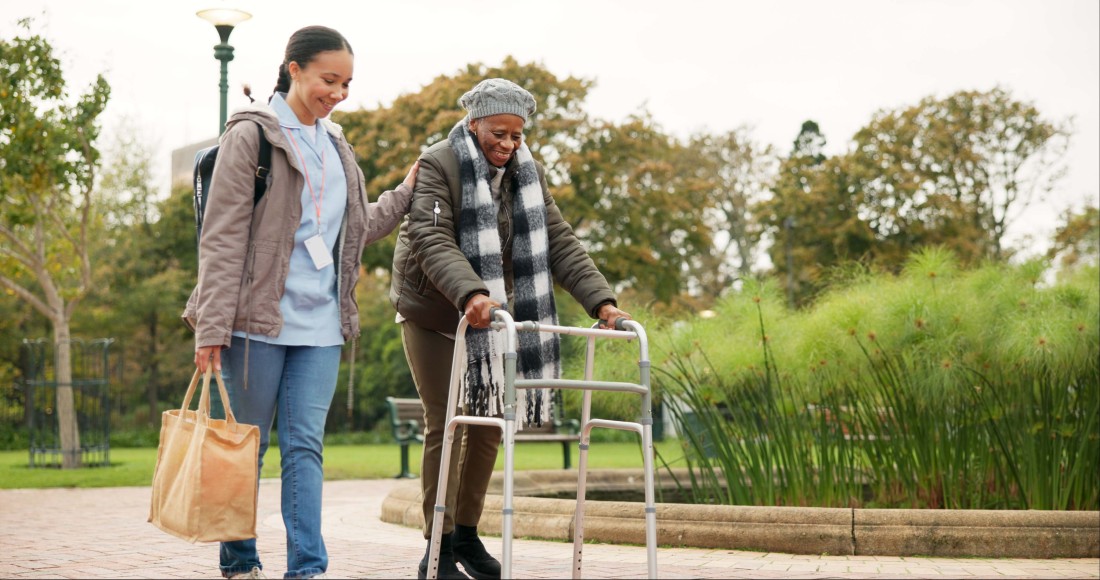 A caregiver carries groceries and accompanies a senior client through the park