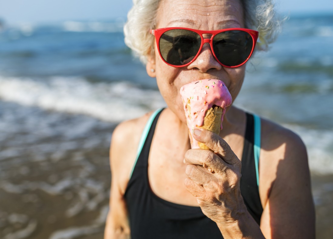 An older woman wearing sunglasses eats ice cream at the beach