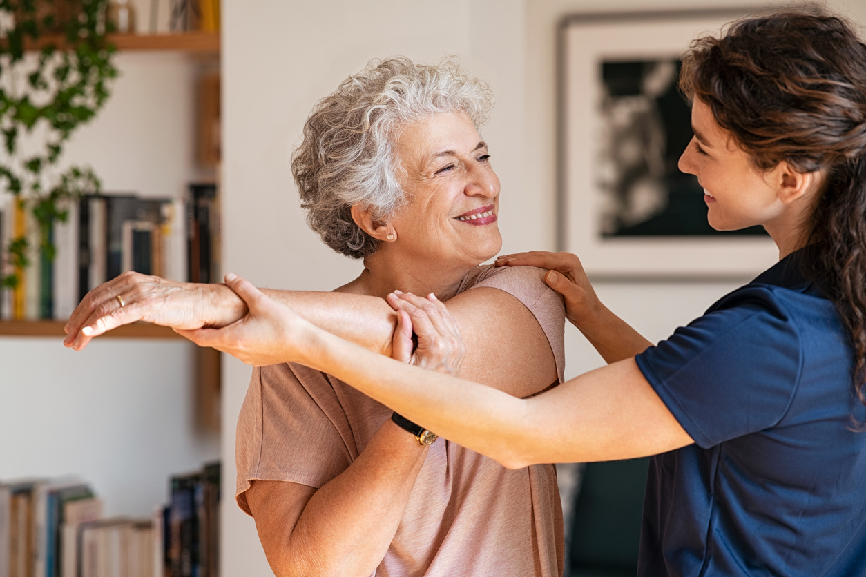 A caregiver helping an older adult with gentle exercises in a bright home living room.