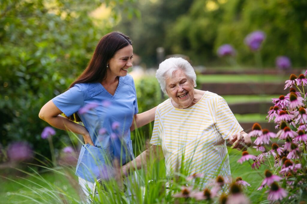 A caregiver walking through the garden with an elderly client, both smiling.